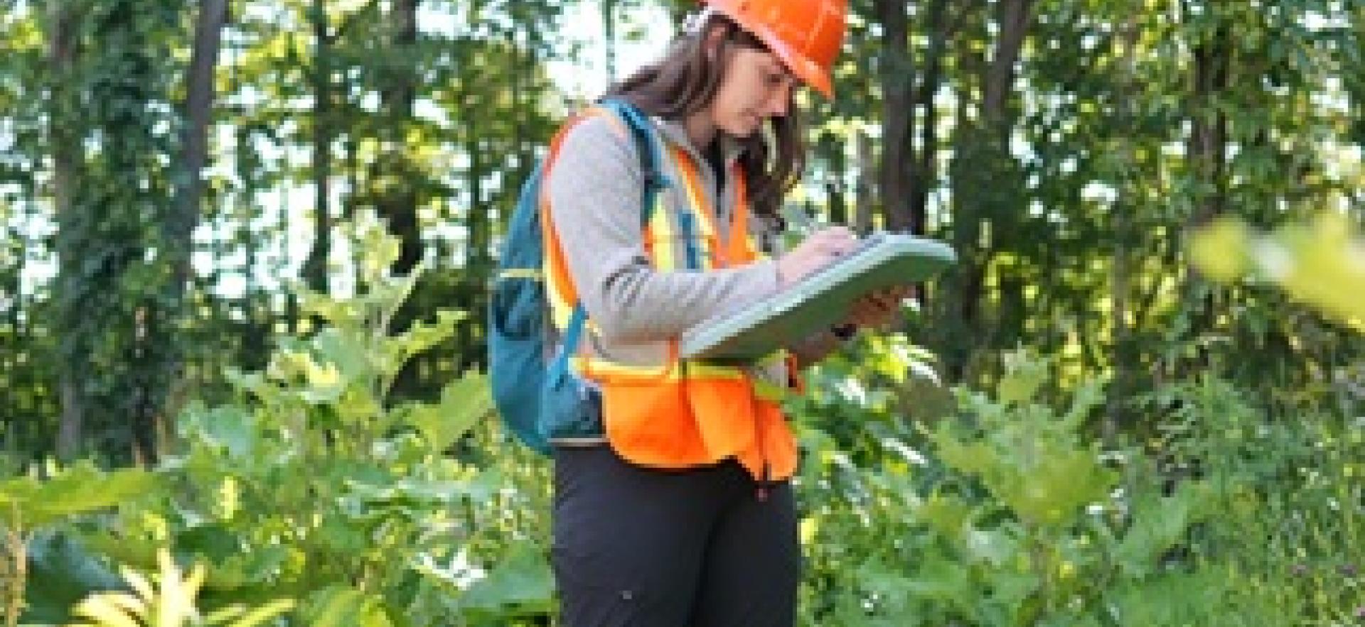 SONE student in the field with hard hat and vest writing down on clipboard