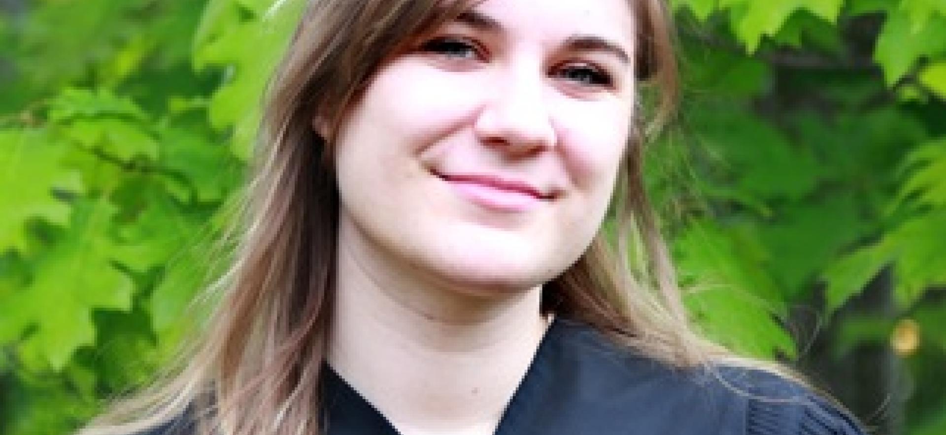 Graduate in gown with trees behind smiling at camera