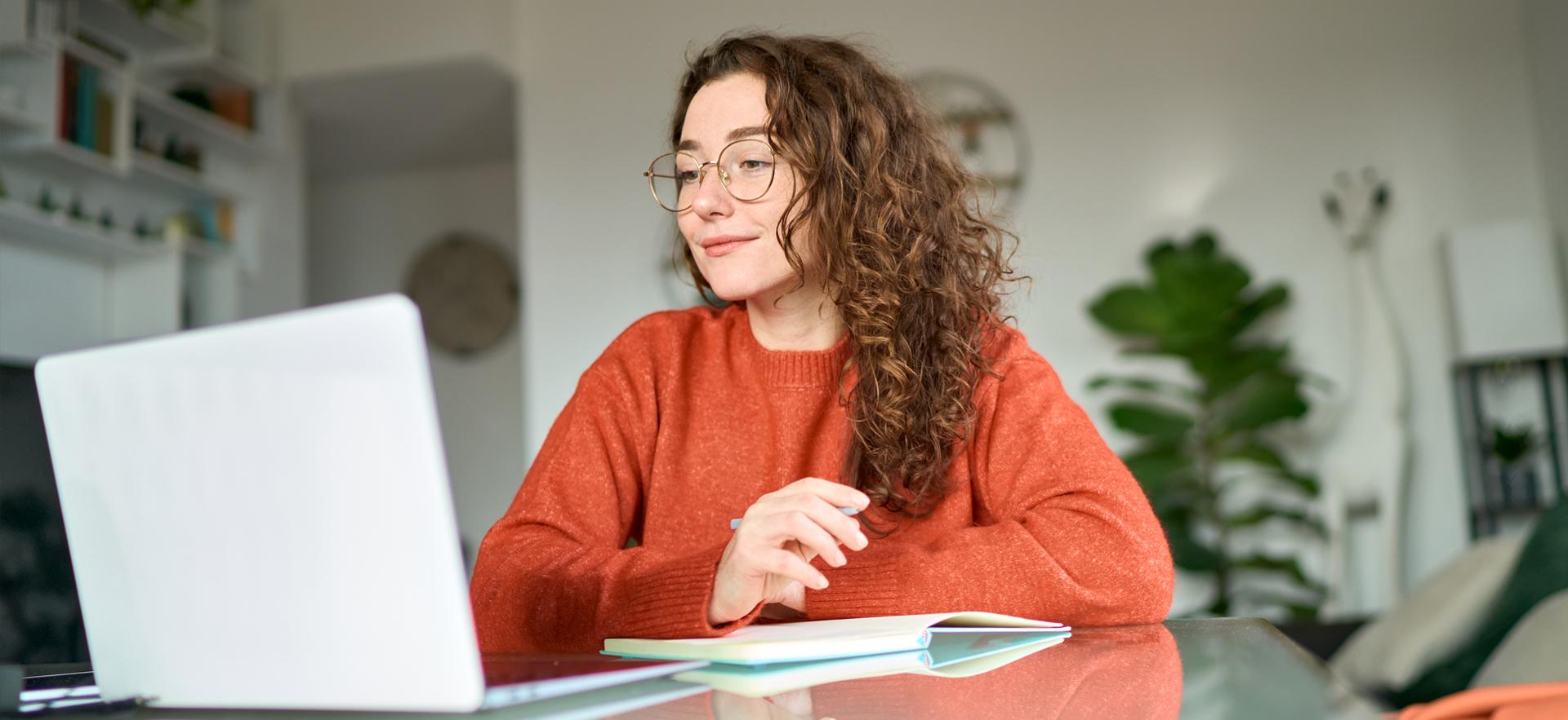 Woman sitting at table looking at laptop with living room in background