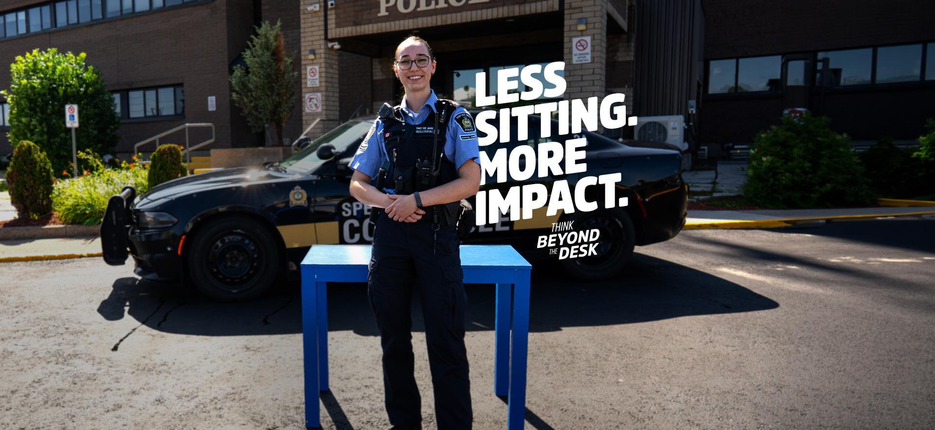 Policing graduate standing in front of blue desk with cruiser parked in front of station