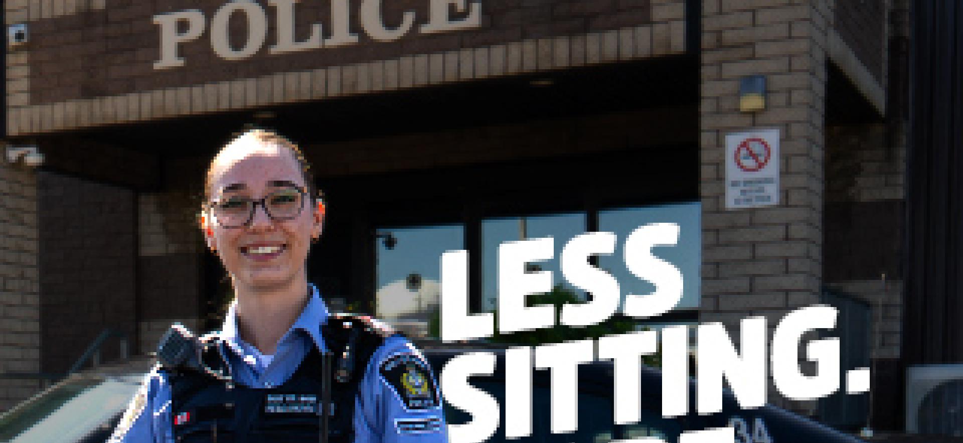 Policing graduate standing in front of blue desk with cruiser parked in front of station
