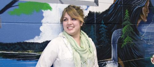 Female indigenous studies student smiling in front of mural.